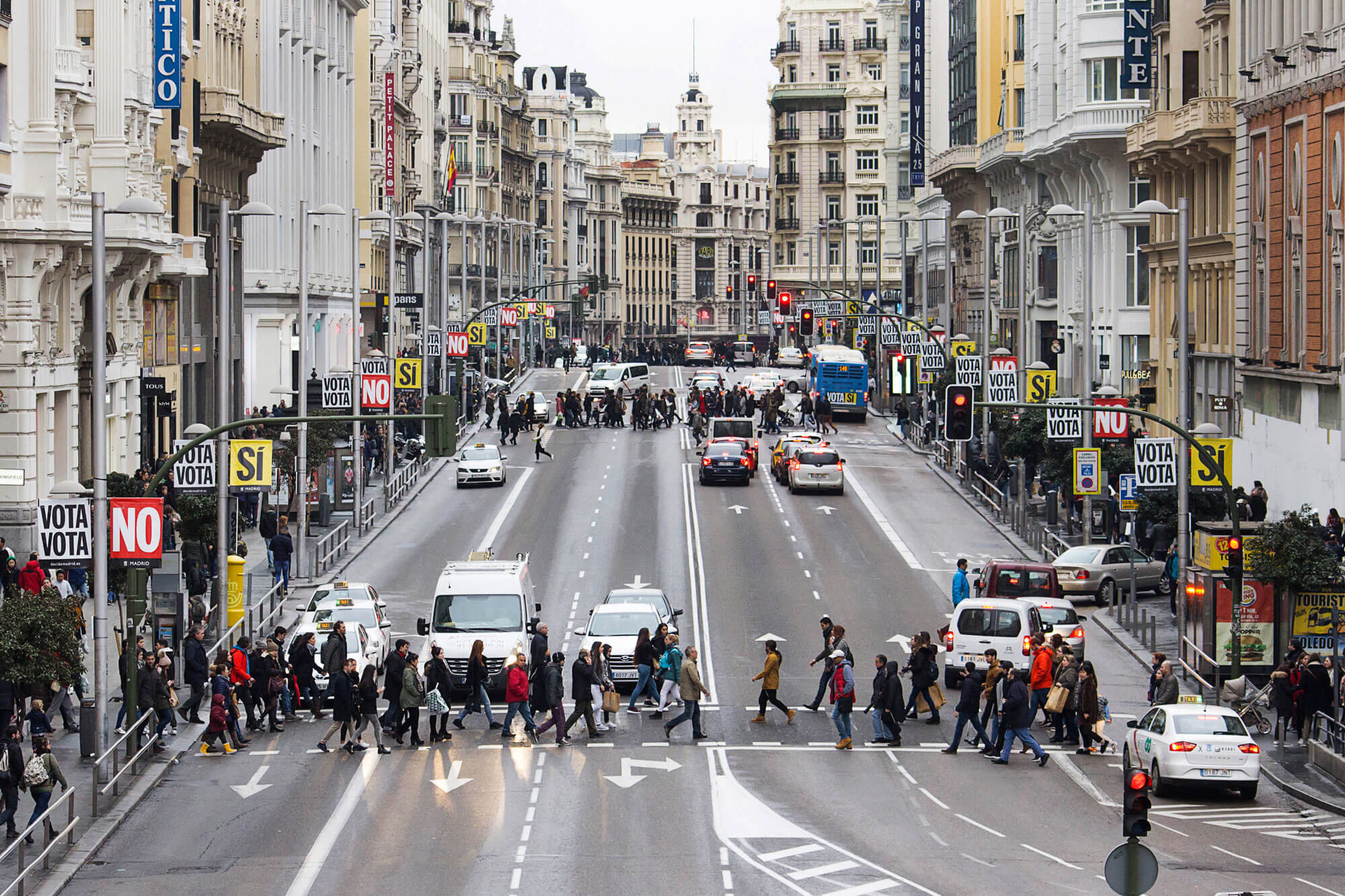Vista panorámica de la calle Gran Vía donde se ven dos cruces de peatones con numerosas personas cruzando. A los lados de la calle hay banderolas en las farolas con la campaña de Decide Madrid vota si vota no