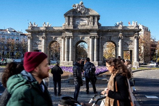 Un hombre con gorro y una mujer con un carrito en primer plano cruzan un paso de cebra con la Puerta de Alcalá de fondo