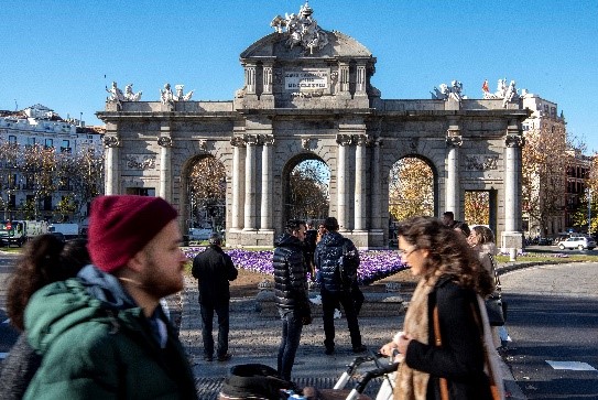 Un hombre con gorro y una mujer con un carrito en primer plano cruzan un paso de cebra con la Puerta de Alcalá de fondo