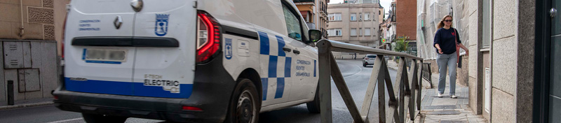 furgoneta del Ayuntamiento de Madrid circula por una calle estrecha con barandillas metálicas, mientras una mujer camina por una acera reducida junto a los edificios.